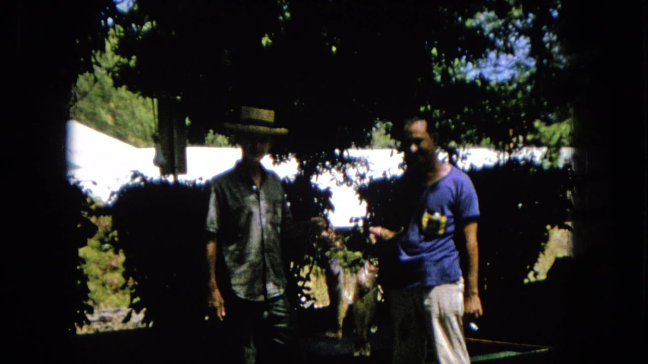 A shaded 1965 home-movie frame shows two adult men outdoors, one in a straw hat and patterned shirt and the other in a blue t-shirt.