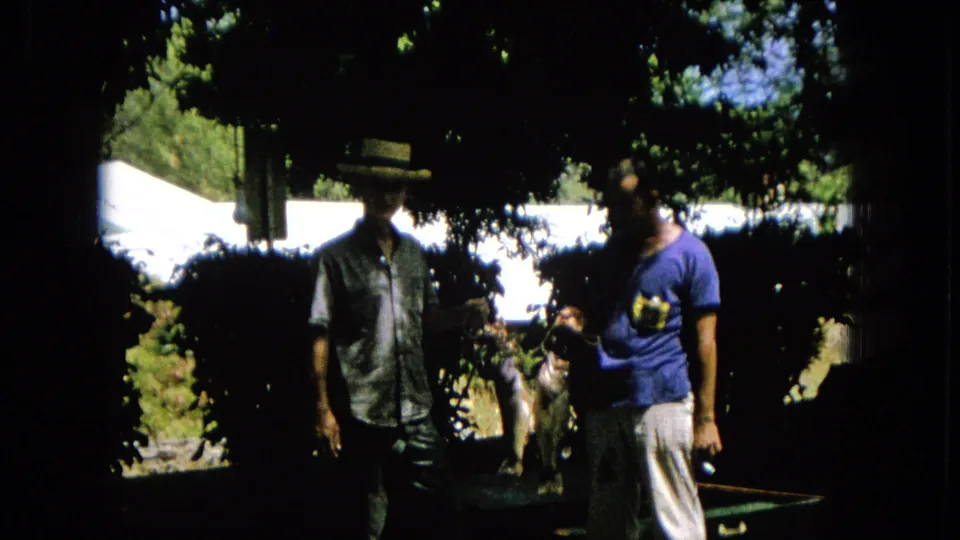 1965 8mm footage frame of two men standing in a shaded outdoor area, with a hanging animal-like form visible near the center.