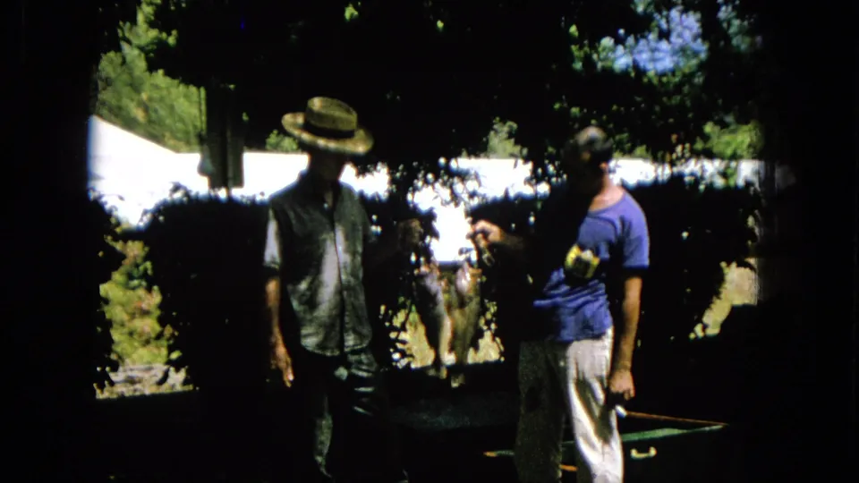 A 1965 outdoor home-movie frame shows two adult men beneath leafy shade, one gesturing beside a hanging animal-like form.