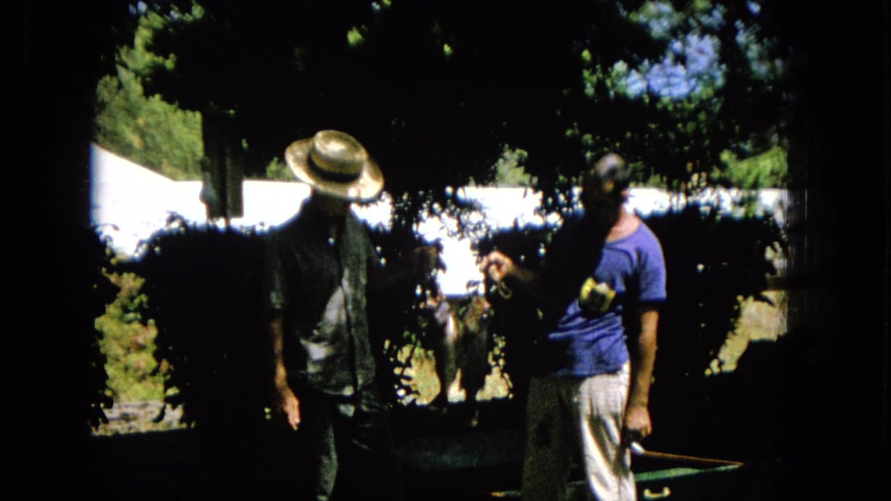 Two adult men stand in a shaded United States garden in 1965 home-movie footage, with one man in a straw hat and patterned shirt and the other in a blue T-shirt.