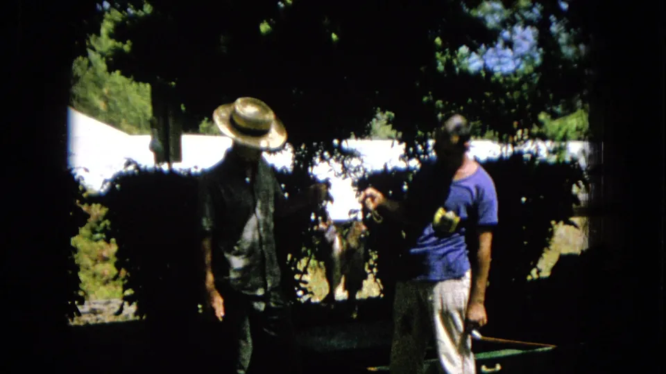 Two adult men stand in a shaded United States garden in 1965 home-movie footage, with one man in a straw hat and patterned shirt and the other in a blue T-shirt.
