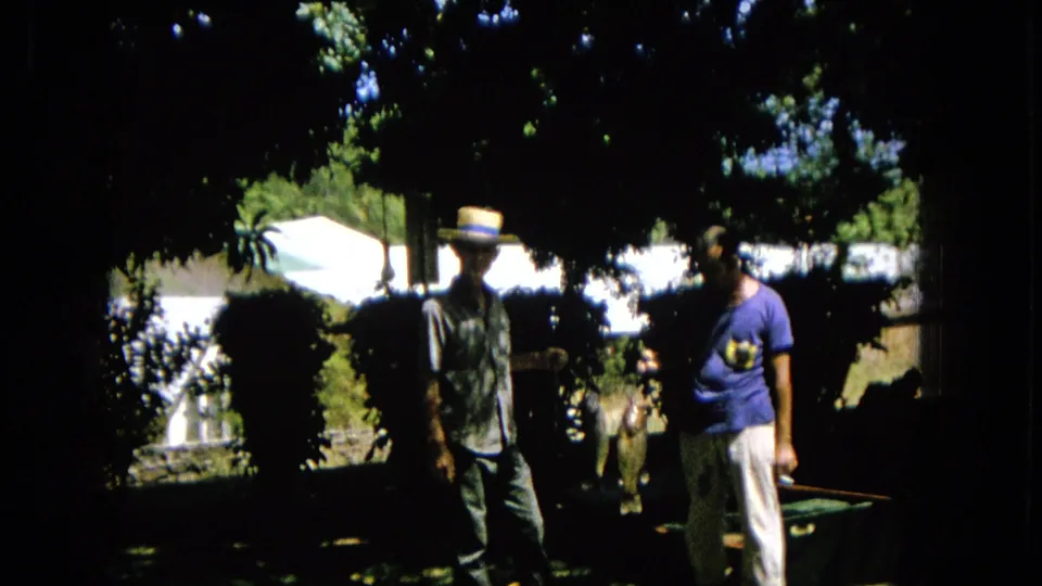 In a dark, sun-dappled garden frame, two adult men appear to converse, with leafy branches overhead and a white wall or building visible in the background.