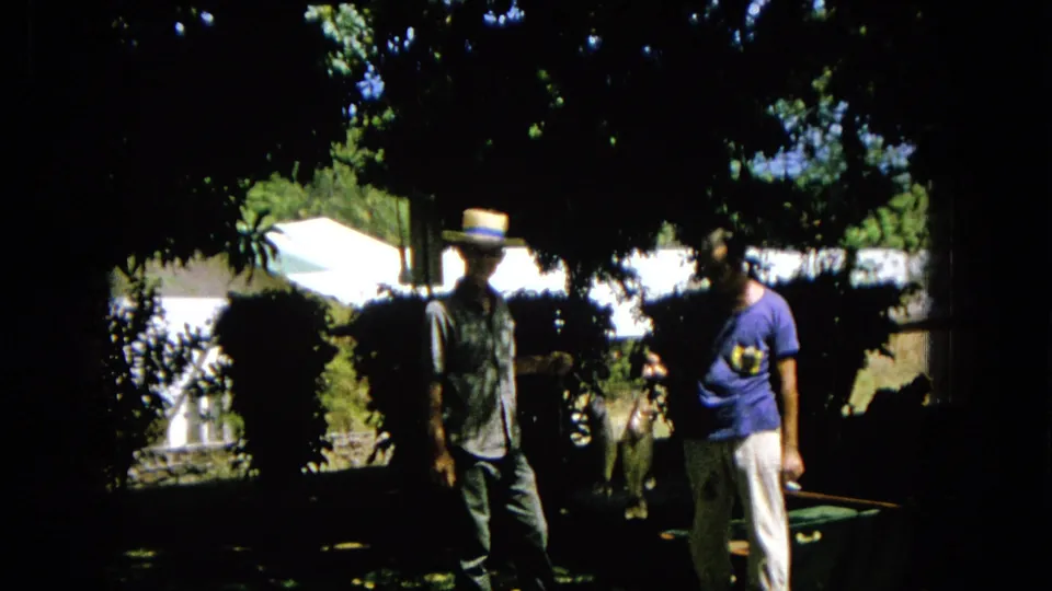 A 1965 home-movie frame shows two adult men in a garden, partly obscured by shadow, with the straw-hatted man standing closer to the camera.