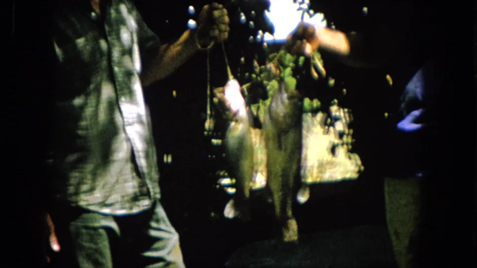 A close Super 8 frame shows two freshly caught fish, likely bass, suspended from strings as the men display them in the shaded backyard.