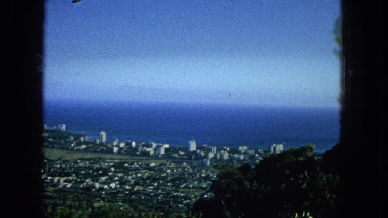 1965 Super 8 frame showing an elevated Hawaiian coastal city panorama with a blue ocean horizon, dense urban blocks, shoreline buildings, and dark vegetation at the lower edge.