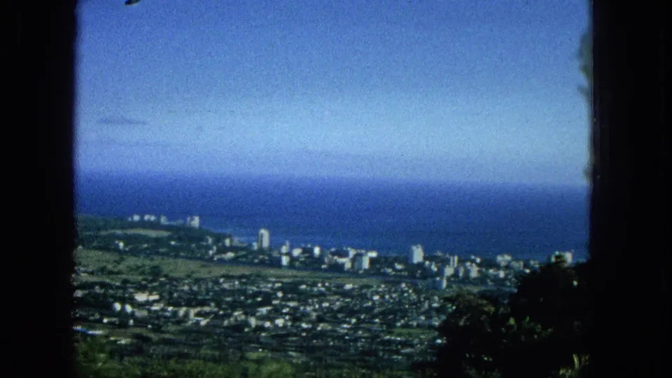 Elevated 1965 Super 8 view across a Hawaiian coastal city, with mid-century shoreline buildings, a broad blue ocean, pale sky, and vegetation framing the lower right.