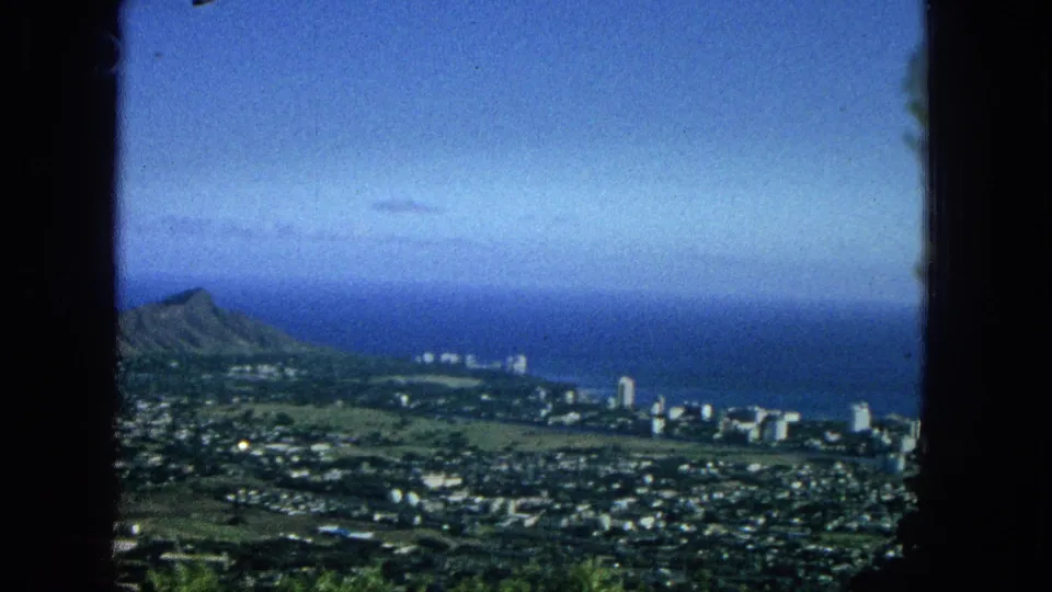 1965 Super 8 frame showing Diamond Head in the distance beyond a coastal urban area, with ocean, beachfront buildings, green land, and a soft blue sky.
