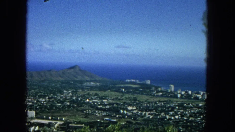 Elevated 1965 Super 8 view of Diamond Head with the Hawaiian coastline, blue ocean, beachfront high-rises, dense city blocks, and green vegetation in the foreground.