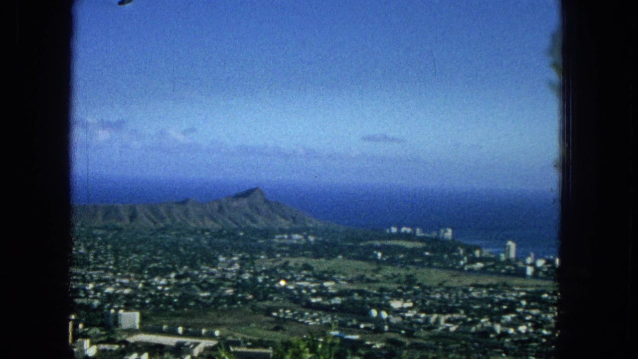 1965 Hawaii home-movie frame showing Diamond Head crater above Honolulu neighborhoods with the Pacific Ocean and blue sky beyond, viewed through dark window edges.