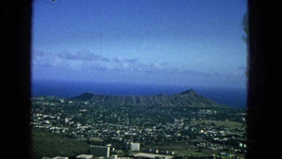 1965 home-movie still showing a softer, slightly lower view across Honolulu toward Diamond Head and the Pacific Ocean, with grain and saturated blue tones visible.