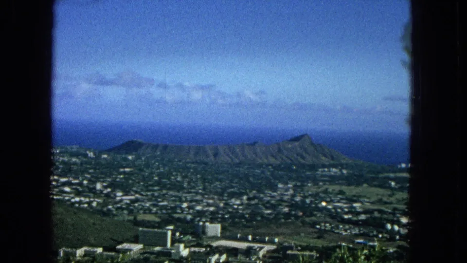 Final 1965 frame looking across Honolulu toward Diamond Head, with city blocks, green land, distant water, and a blue sky framed by dark film or window edges.