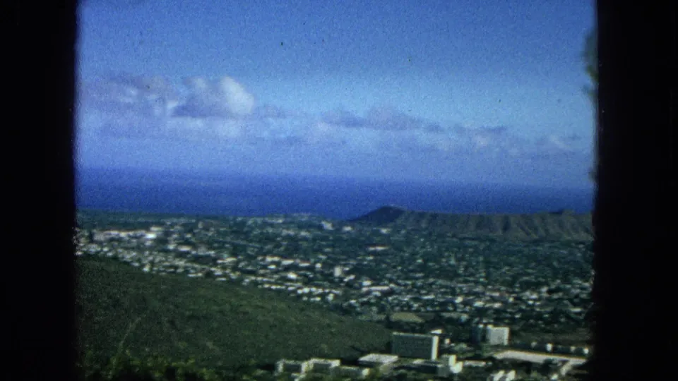 1965 Oahu panoramic frame showing dense low-rise development, green hillside terrain, Diamond Head near the right side, blue ocean, and a dark vertical window divider.
