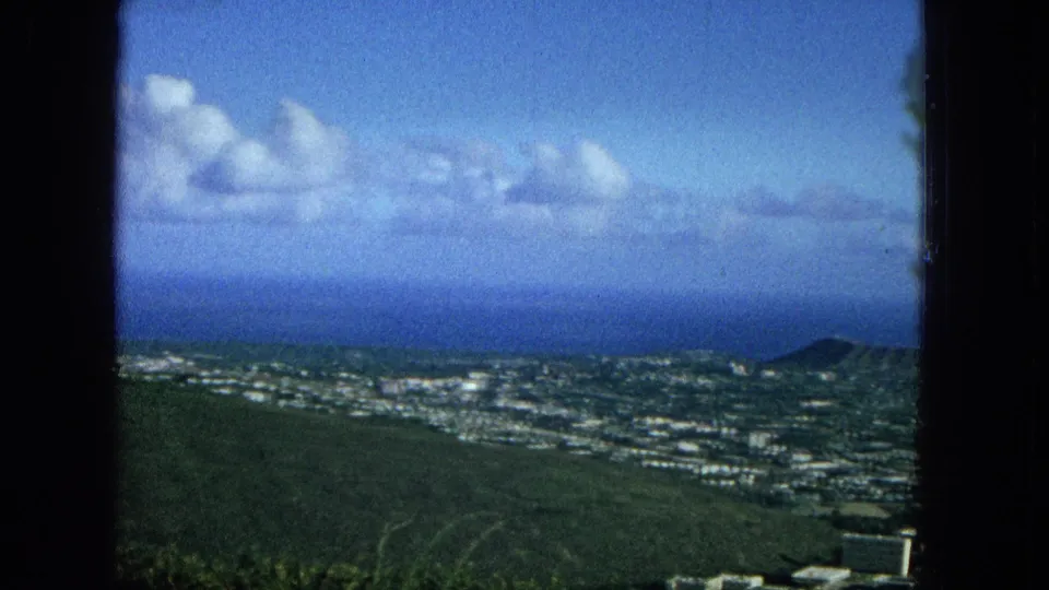 1965 Oahu home-movie frame with a green hillside in the foreground, distant residential and urban areas, ocean horizon, scattered clouds, and a dark window-frame border.