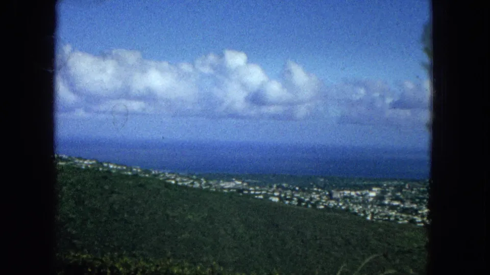 1965 Oahu view across a green hillside toward a spread of city buildings, the Pacific horizon, large white clouds, and dark window-frame borders at the sides.