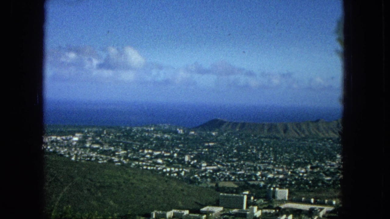 1965 8mm frame looking across Honolulu toward Diamond Head crater and the Pacific Ocean under a partly cloudy sky.