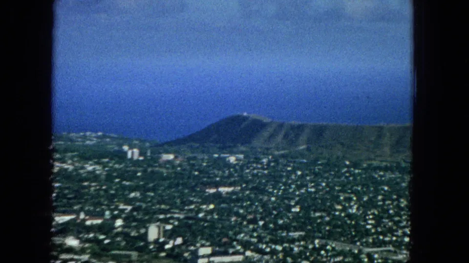 1965 Honolulu cityscape seen from an elevated or aerial viewpoint, with Diamond Head crater rising behind dense neighborhoods and the Pacific Ocean in the distance.