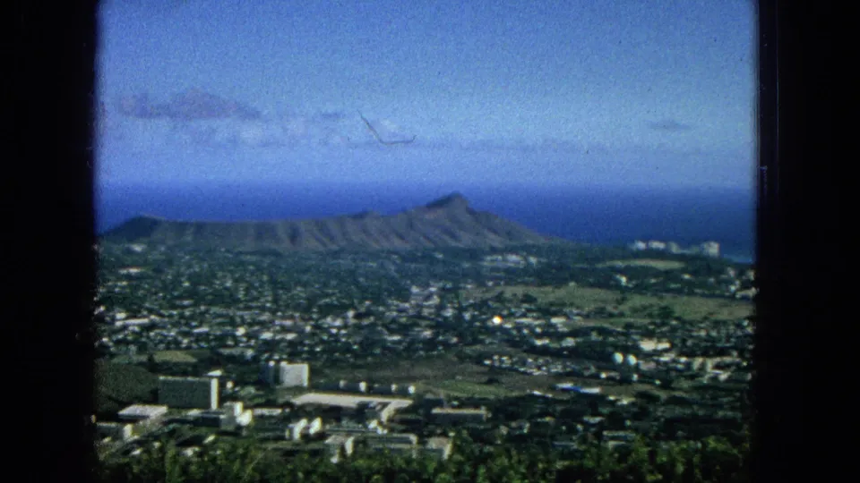 Wide 1965 Honolulu view with Diamond Head stretched across the horizon, low-rise urban development below, greenery in the foreground, and the ocean behind.
