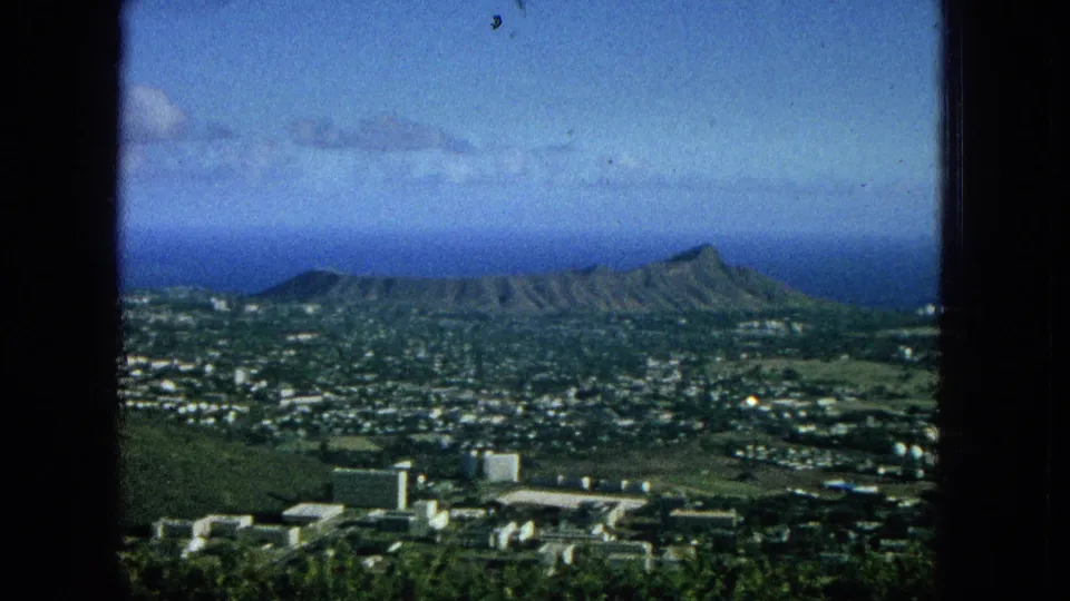 1965 elevated view over Honolulu toward Diamond Head, showing city blocks, green open areas, distant ocean, and a clear blue sky.