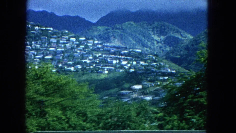 Distant Molokai hillside settlement seen from an elevated viewpoint, with clustered roofs, green slopes, and blue mountain forms in the background.
