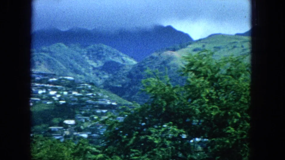 Super 8 frame showing part of a Molokai hillside village at lower left, with green trees and layered mountain slopes filling the view.