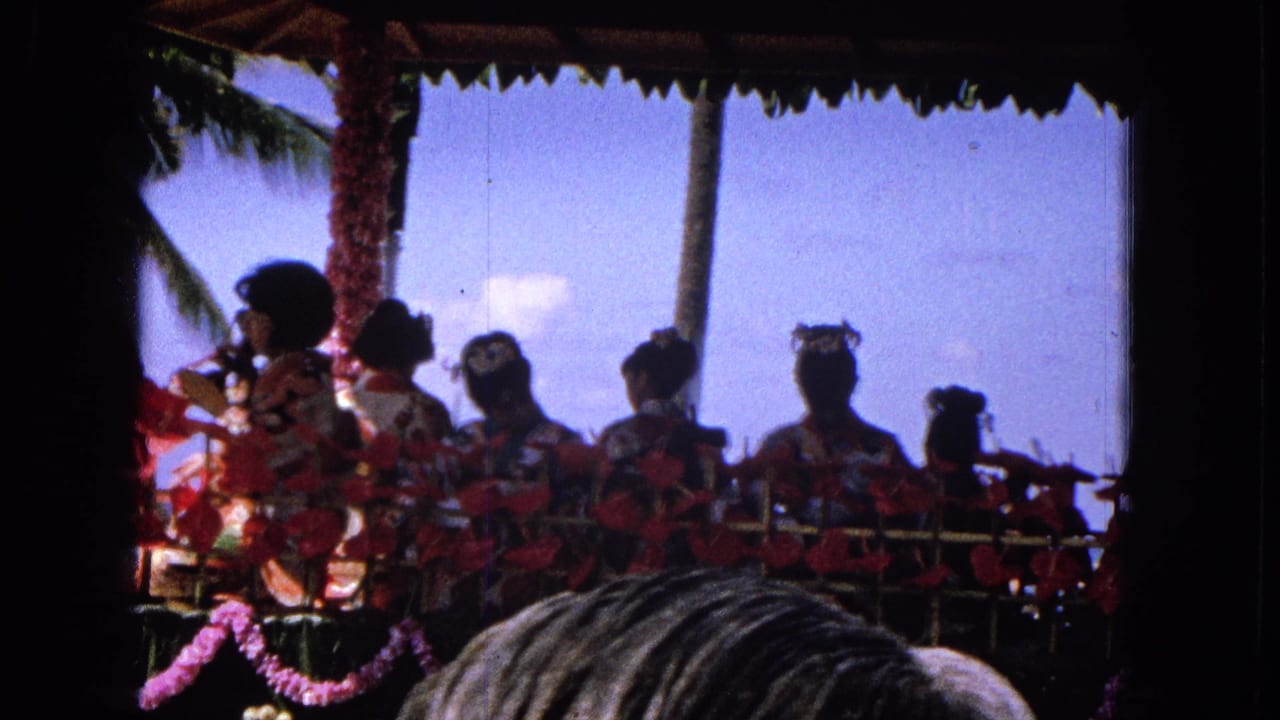 1965 home-movie frame of a Hawaiian parade float with performers in red costumes, flower decorations, palm trees, blue sky, and spectators partially blocking the lower foreground.