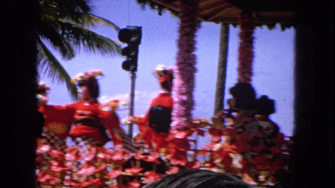 Blurred performers in red costumes on a floral parade float near palm trees and a traffic signal.