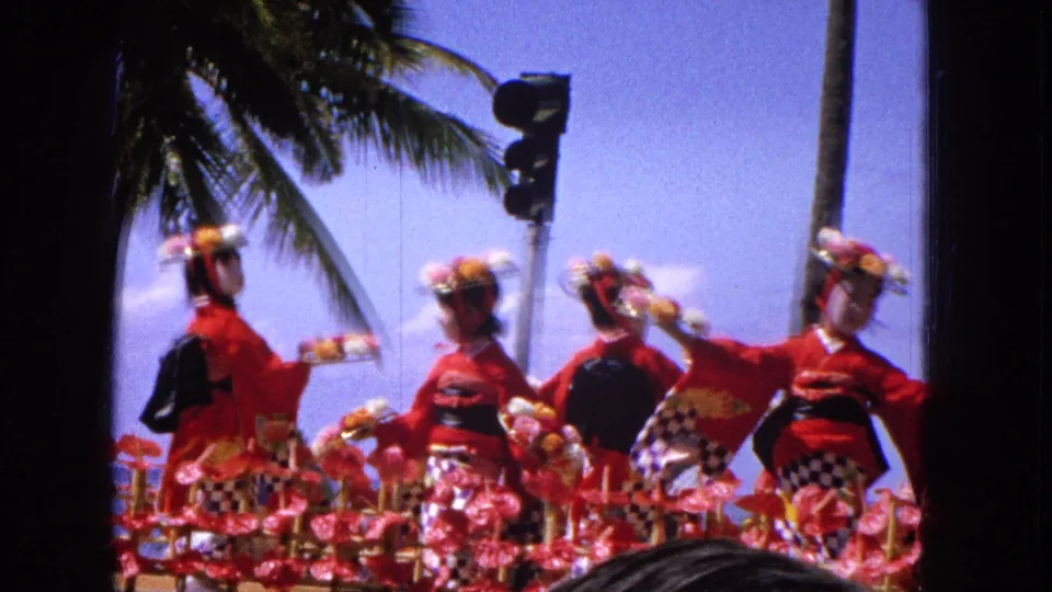 1965 Hawaiian parade footage frame with several performers in red and black geisha-inspired costumes standing on a decorated float under palm trees, with spectators at the bottom of frame.