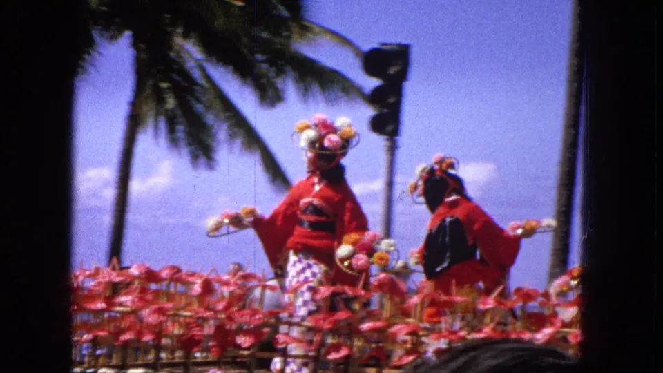 1965 home-movie frame of two performers in red geisha-inspired parade costumes on a floral float, framed by palm trees, blue sky, a traffic signal, spectator heads, and dark film borders.