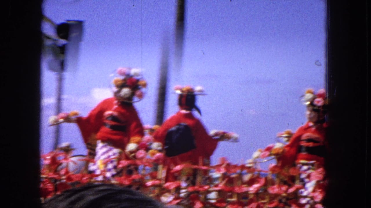 1965 Hawaiian parade frame showing women in red costumes and floral headdresses on a decorated float, viewed from a spectator position with traffic lights nearby.