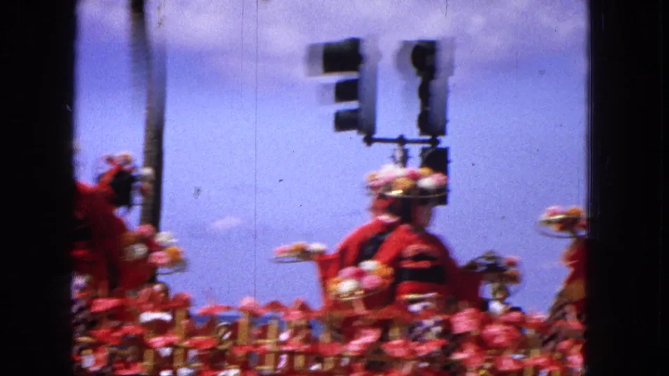 Spectator-shot 1965 parade view with motion-blurred women in red outfits riding a floral float near street traffic lights.