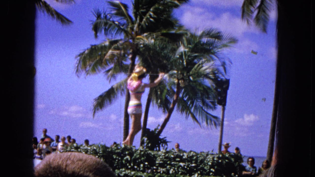 1965 Molokai parade footage shows a performer in a colorful swimsuit on or near a float, with palm trees and beachside onlookers behind her.