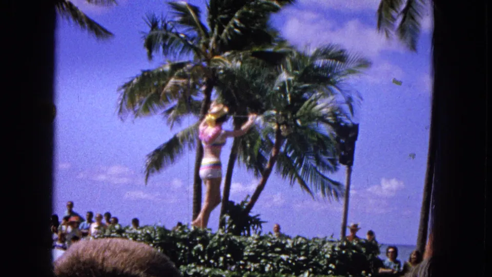 1965 Molokai parade footage shows a performer in a colorful swimsuit on or near a float, with palm trees and beachside onlookers behind her.