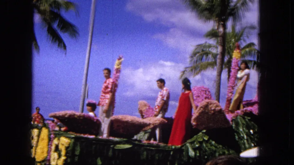 Side view of a decorated floral float in the 1965 Molokai parade, with participants in bright clothing and palm trees rising behind them.