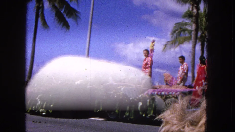 The passing floral parade float is partly obscured by a large white foreground object, with participants, palm trees, and blue sky still visible in the 1965 Molokai scene.
