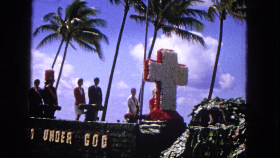 1965 Molokai parade float with a large floral cross, suited participants, women in red jackets, palm trees, and visible "UNITED UNDER GOD" lettering.
