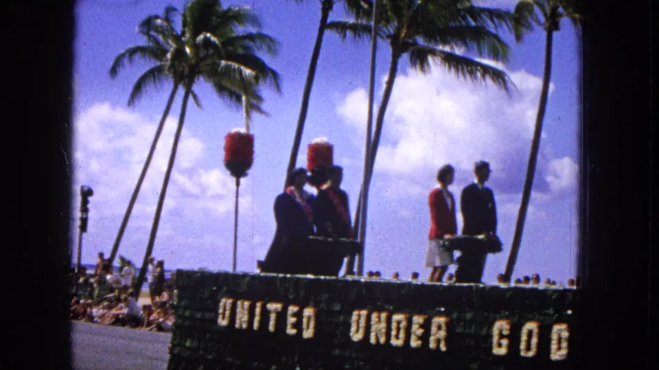 1965 Molokai parade footage shows suited men and women in red jackets on the decorated float, with spectators along the roadside and palm trees behind them.