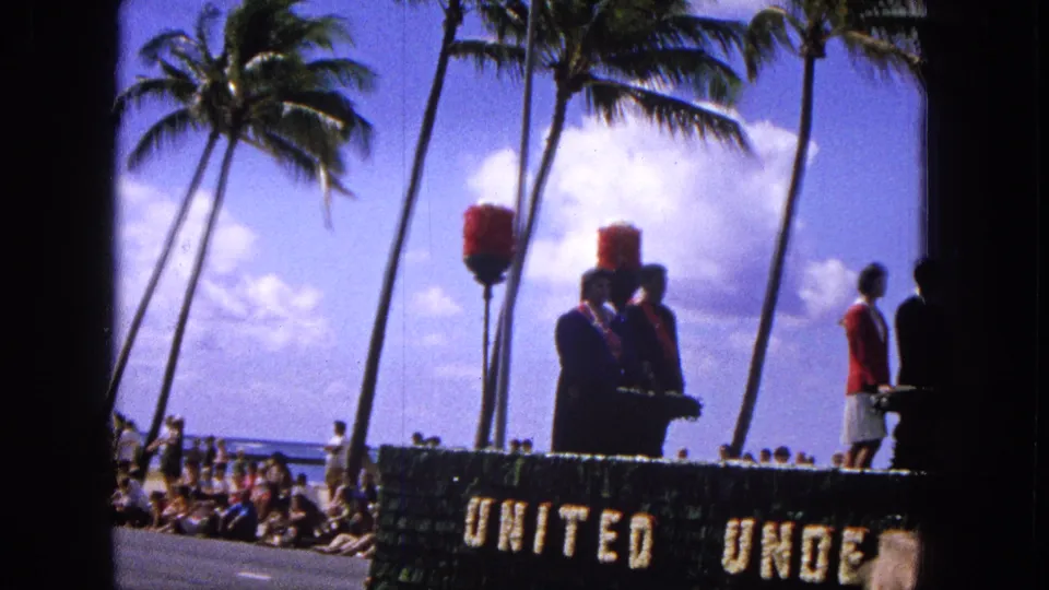 1965 Molokai Easter parade frame with float participants in formal clothing, partial "UNITED UNDER" lettering, palm trees, and seated spectators near the road.
