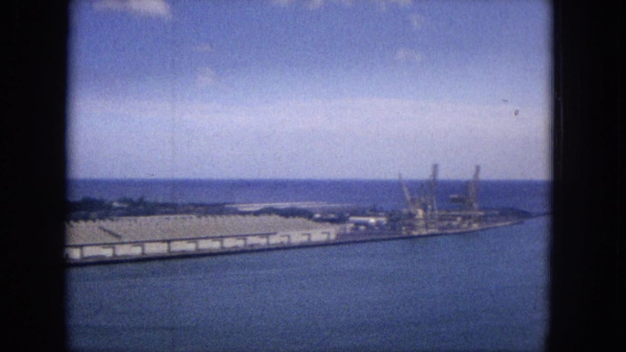 1965 Super 8 view across calm Hawaiian harbor water toward long warehouse-like structures, cranes, shoreline vegetation, and the ocean horizon.