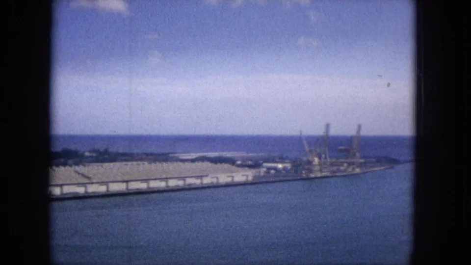 1965 Super 8 view across calm Hawaiian harbor water toward long warehouse-like structures, cranes, shoreline vegetation, and the ocean horizon.
