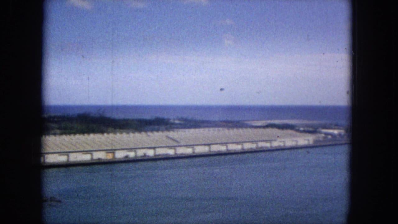 1965 Super 8 frame showing a long Hawaiian coastal pier or breakwater lined with low warehouse-like structures, with blue harbor water in front and the ocean beyond.