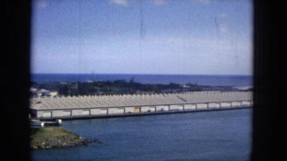1965 Super 8 view of a Hawaiian harbor, with blue water, a small rocky point, long waterfront sheds, distant shoreline, and soft faded color.