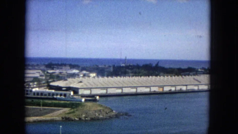 1965 Super 8 frame looking across Hawaiian coastal water toward white-roofed port buildings, distant cranes or masts, shoreline vegetation, and a rocky foreground point.