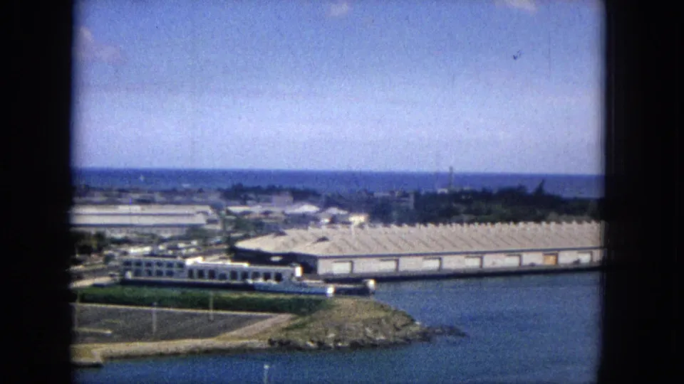 1965 home-movie frame looking across a Honolulu-area waterfront with low industrial buildings, a road network, water, and a bright sky.