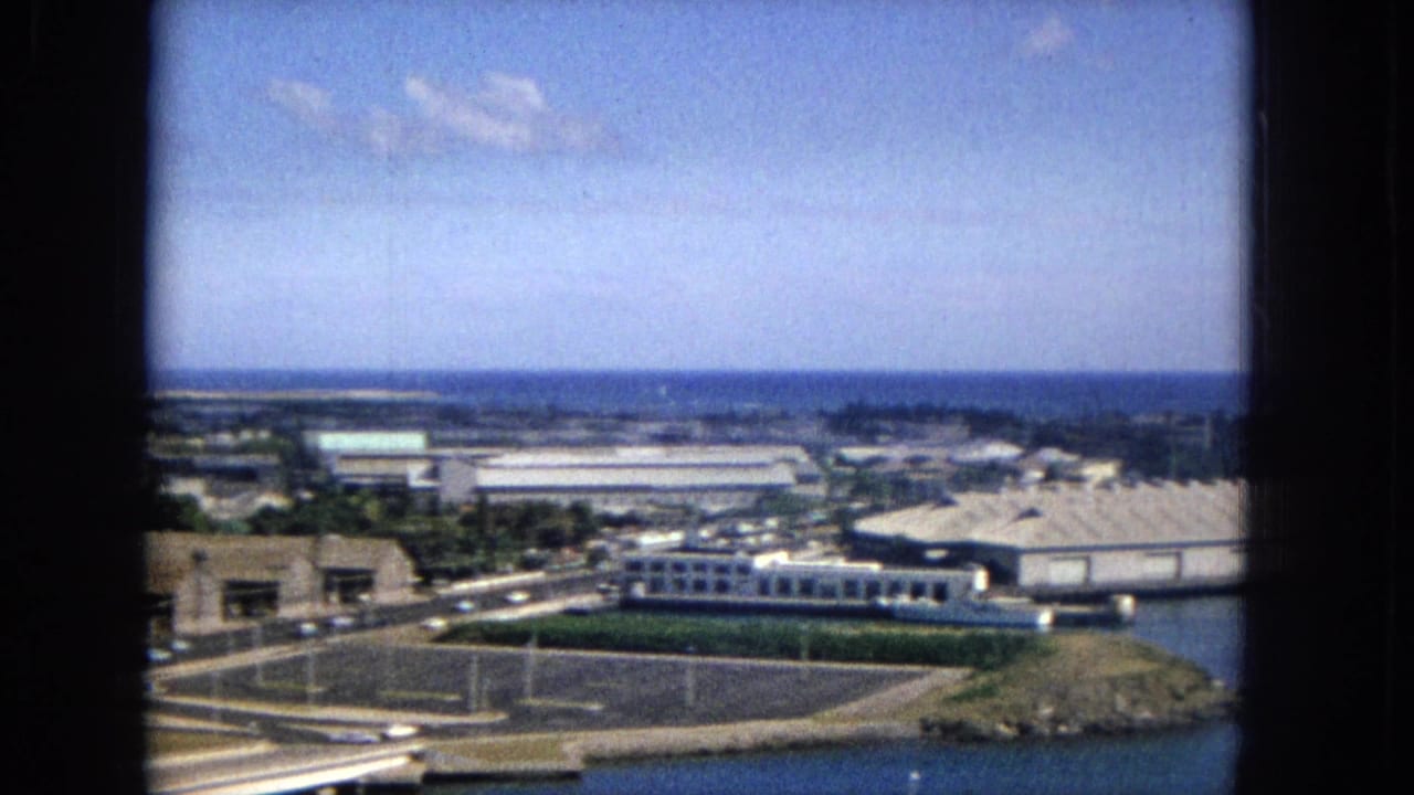 Elevated 1965 view across a coastal Honolulu-area district with warehouse-like buildings, light roadway activity, water, and distant coastline.