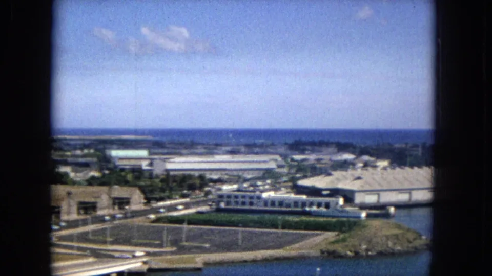 Elevated 1965 view across a coastal Honolulu-area district with warehouse-like buildings, light roadway activity, water, and distant coastline.