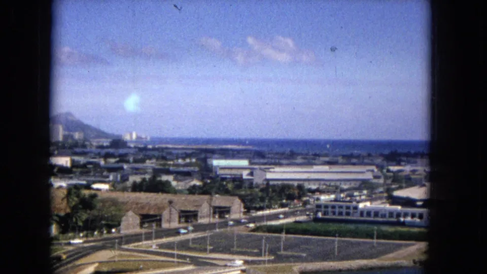 1965 home-movie frame showing a broad Honolulu-area coastal view with roads, low buildings, harbor edge, and Diamond Head visible in the distance.