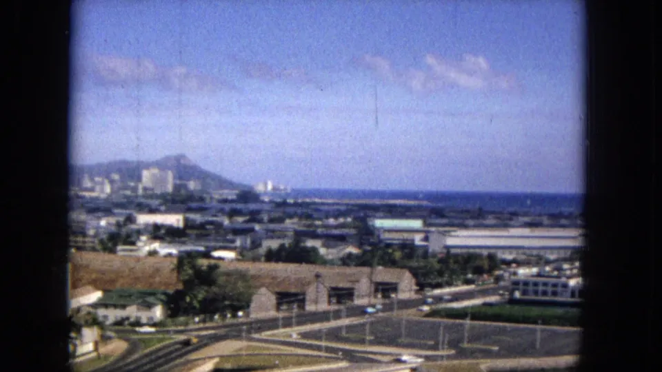 1965 home-movie frame looking over a Honolulu-area waterfront district toward Diamond Head, with roads, low buildings, and the Pacific horizon visible.