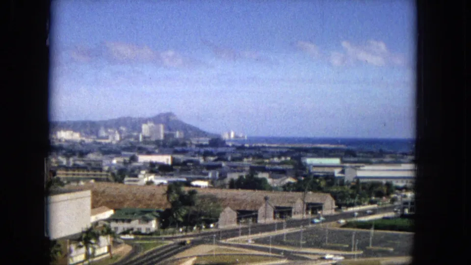 1965 home-movie frame showing an elevated Honolulu overview with Diamond Head beyond mid-century buildings, roads, palm trees, and a bright coastal sky.