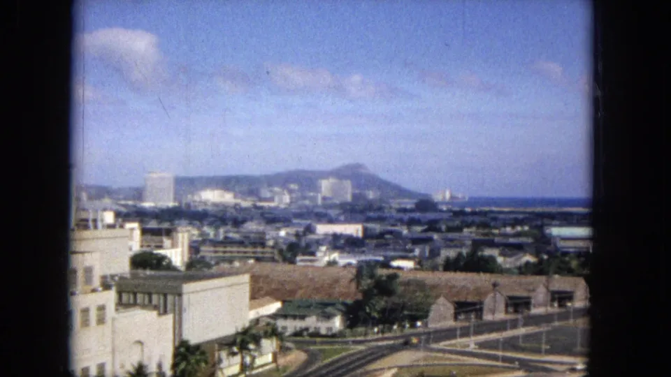 1965 Honolulu city overlook with Diamond Head on the horizon, scattered mid-century buildings, foreground streets, and dark scan borders around the frame.