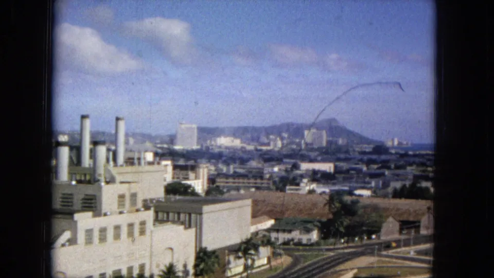 1965 home-movie view across a foreground building and curving roads toward Honolulu towers, distant ridge lines, and the Diamond Head area under scattered clouds.